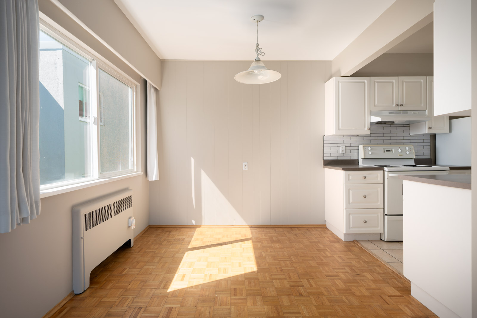 Bright, empty kitchen and dining area with parquet flooring, white cabinets, tiled backsplash, large window, and hanging light fixture.