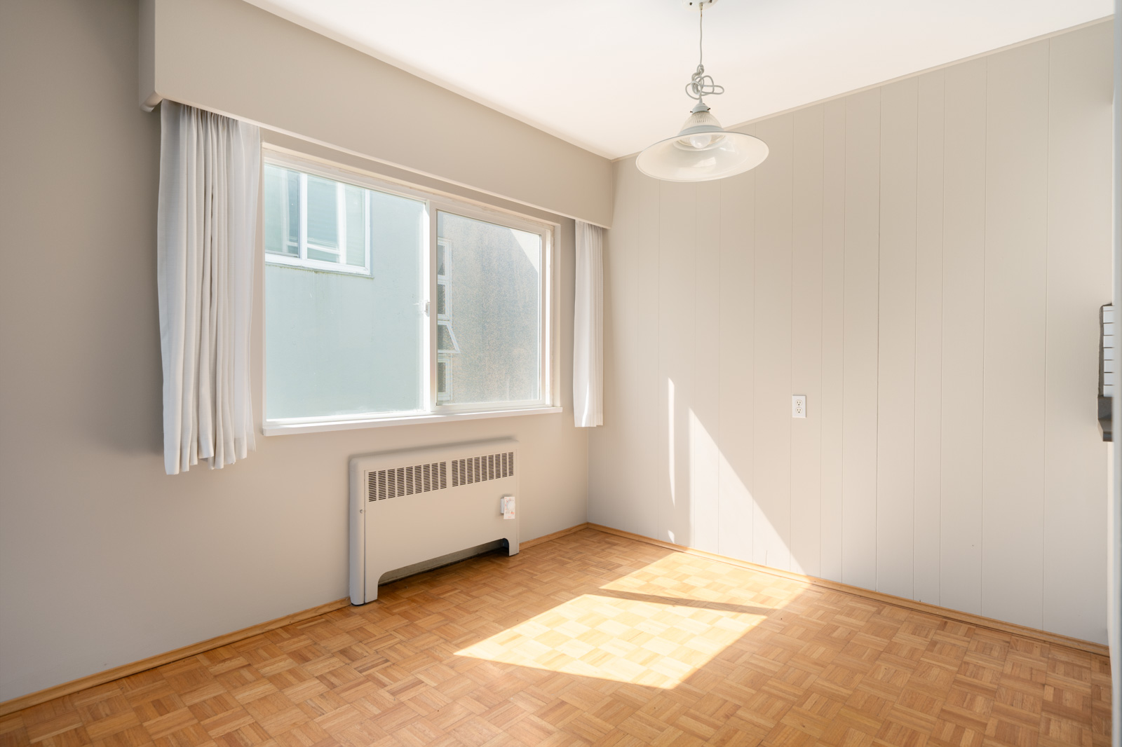 Bright, empty room with parquet flooring, large window with white curtains, a wall radiator, and a ceiling light fixture. Sunlight streams in, illuminating the floor.