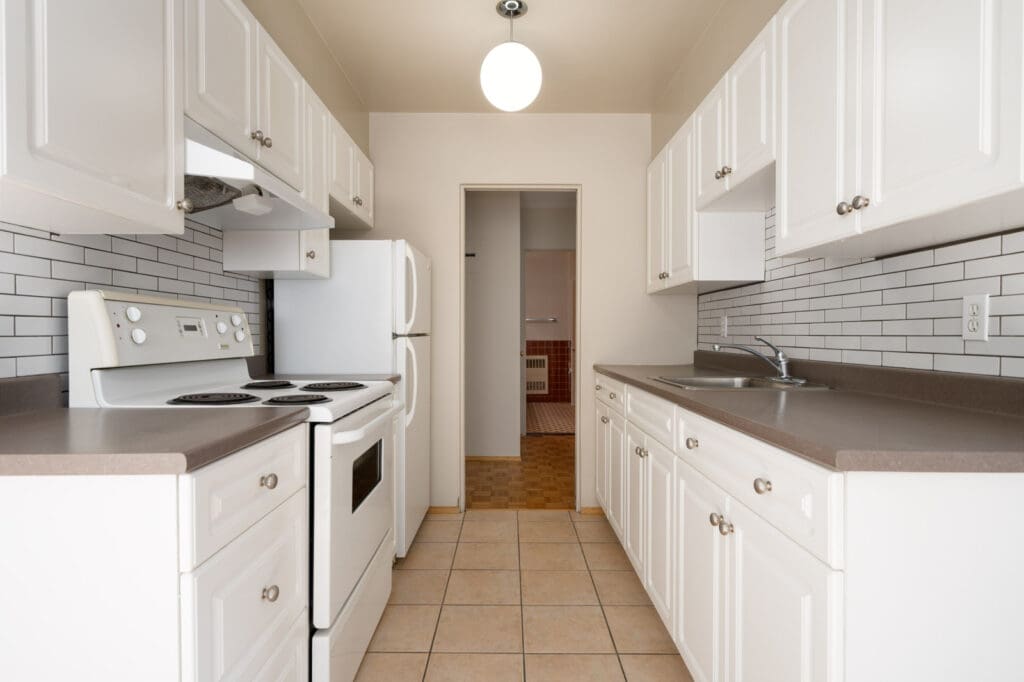 A narrow kitchen with white cabinets, white appliances, brown countertops, tile flooring, and a white tile backsplash. A door at the end leads to another room.