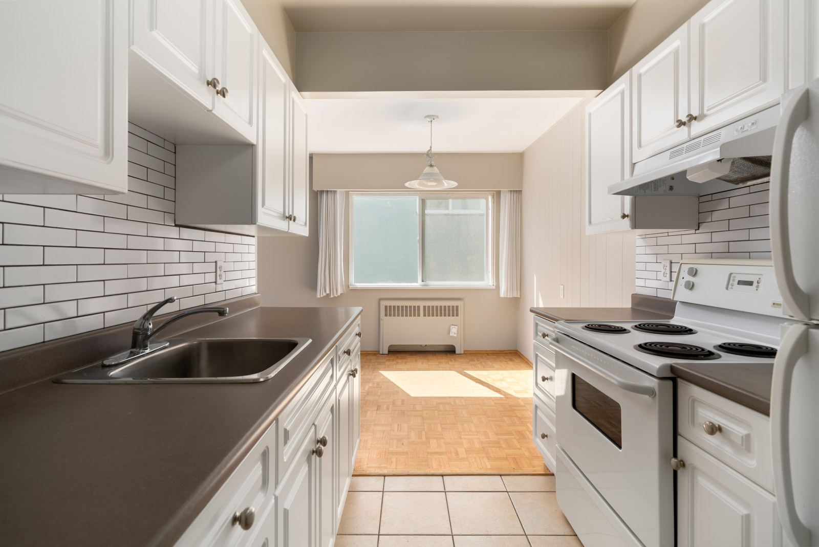 A galley kitchen with white cabinets, tiled backsplash, electric stove, and sink, opening into a dining area with large window and hardwood floor.