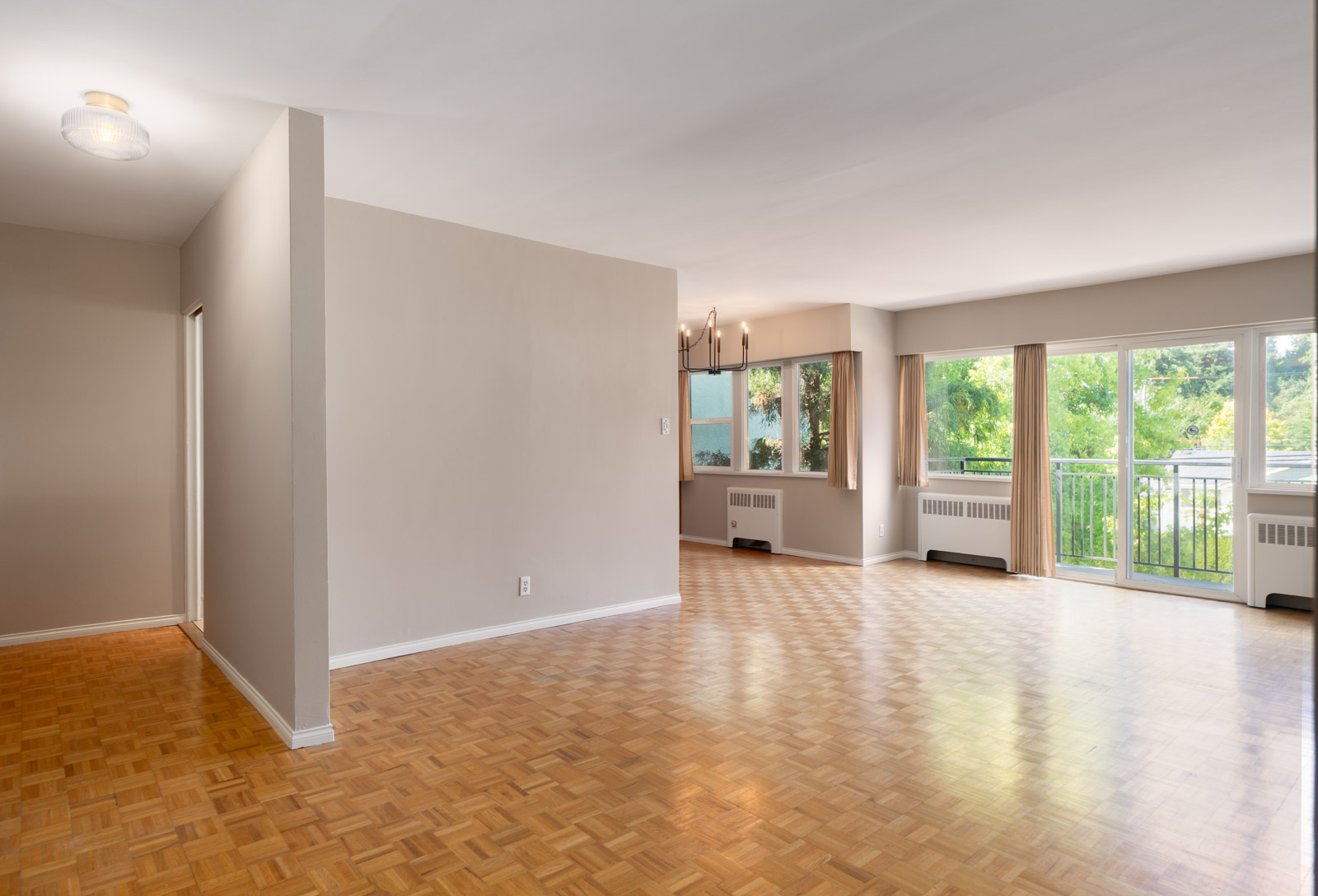 Empty apartment living room with light wood parquet flooring, beige walls, large windows, sliding glass door, and a balcony with a view of greenery.