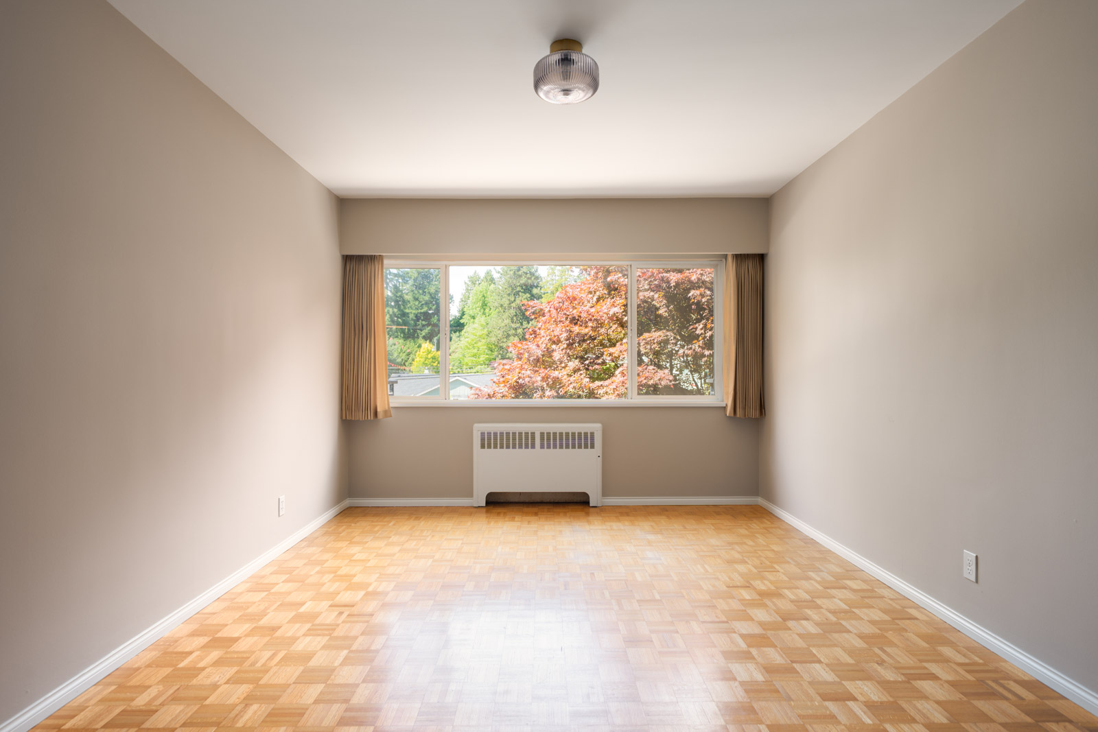 Empty room with light-colored parquet flooring, beige walls, a ceiling light, large window with tan curtains, and a view of trees outside.