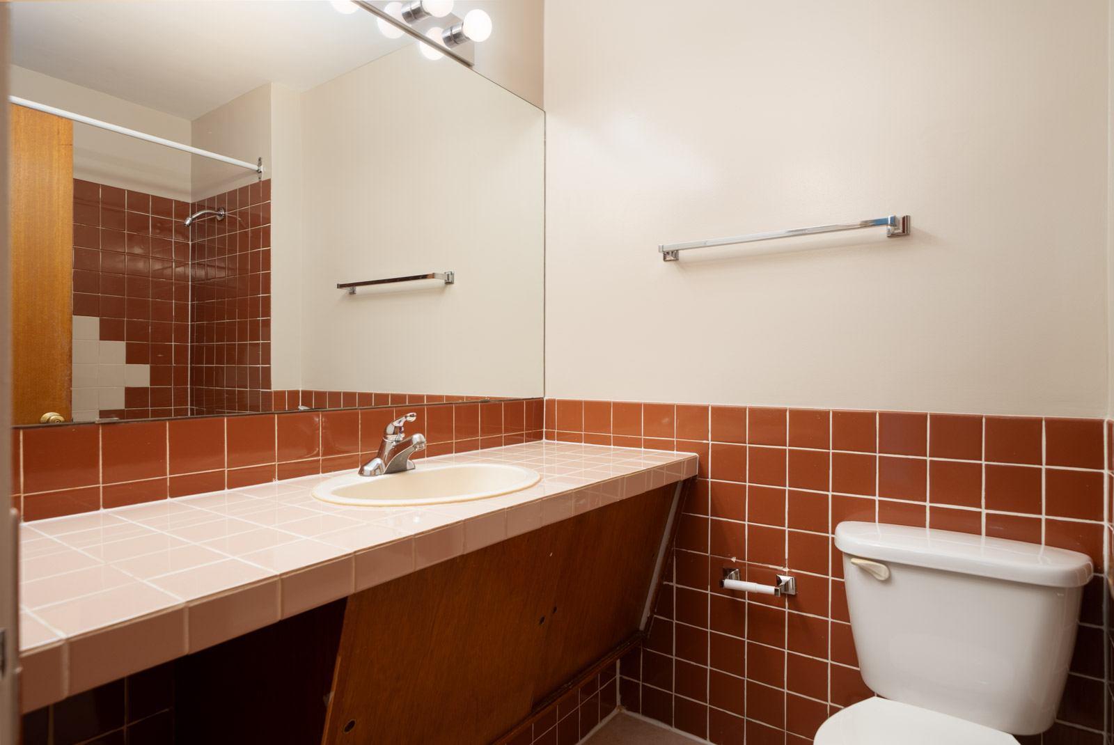 Bathroom with a tiled countertop and sink, red-brown wall tiles, a toilet, a towel bar, and a large mirror above the sink.