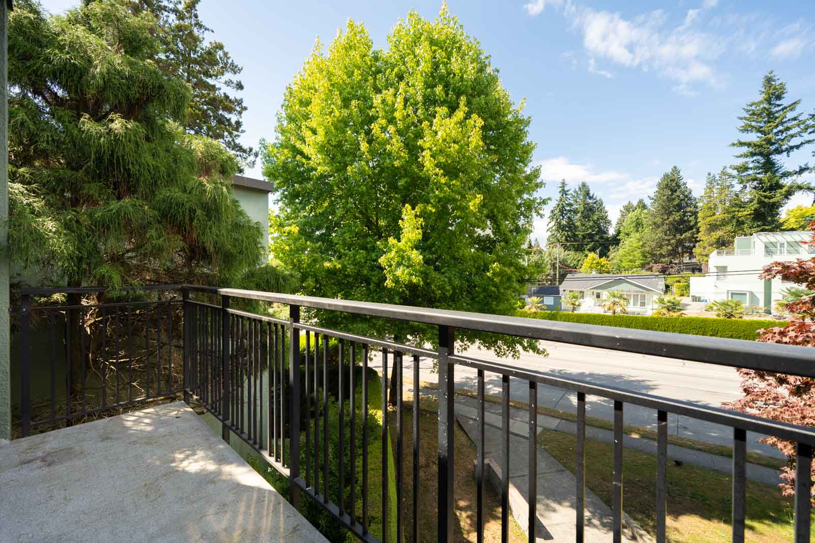 View from a balcony with a black metal railing, overlooking a tree-lined residential street on a sunny day.