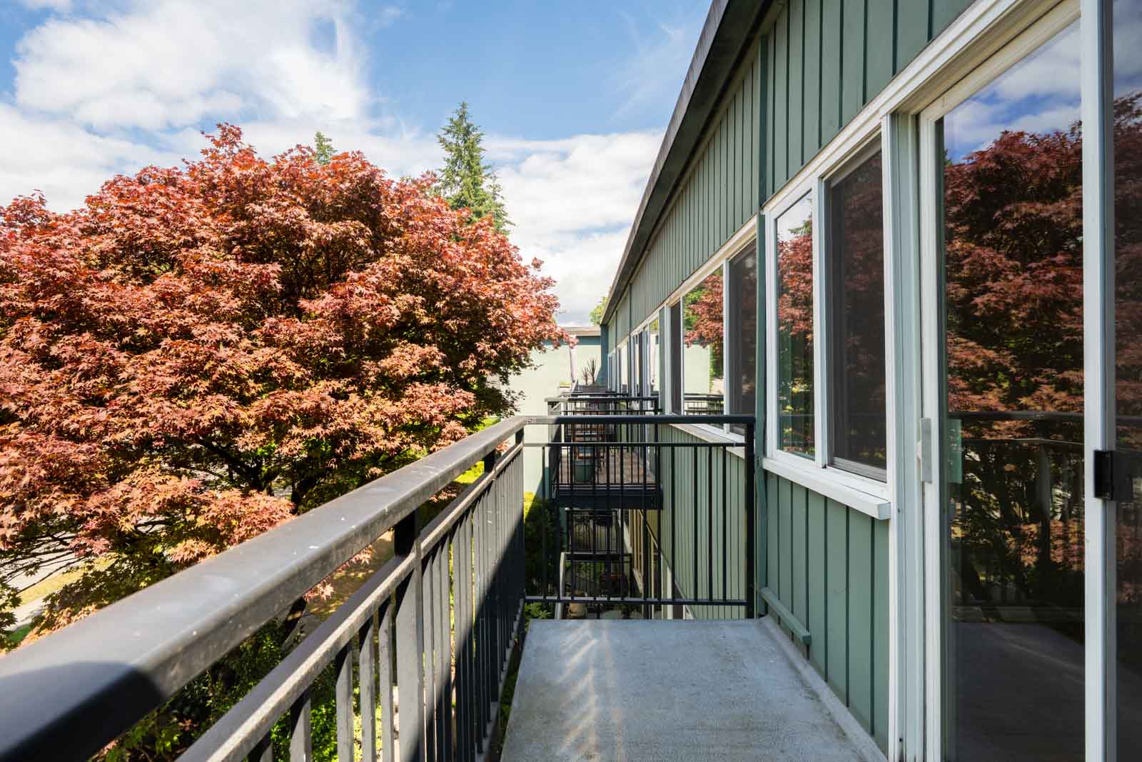 A narrow apartment balcony with metal railings overlooks a tree with red leaves and the building’s green exterior siding under a partly cloudy sky.