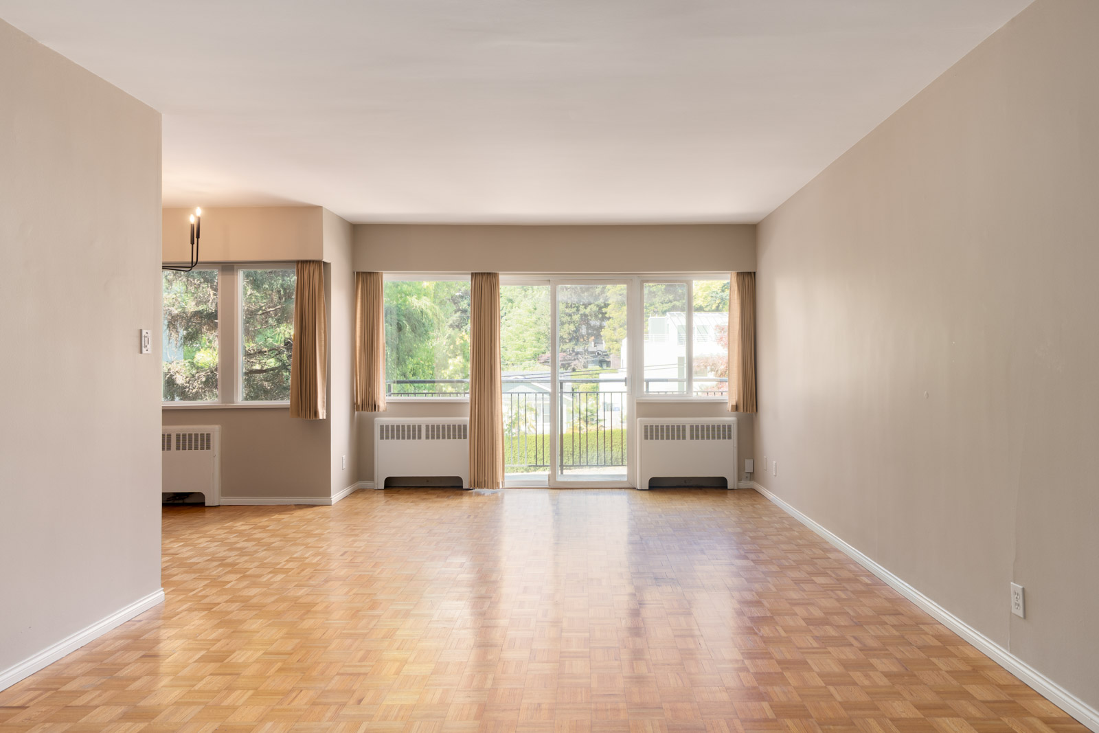 Unfurnished living room with parquet floors, large windows, beige walls, and a sliding glass door leading to a balcony. Dining area with a hanging light fixture is visible on the left.