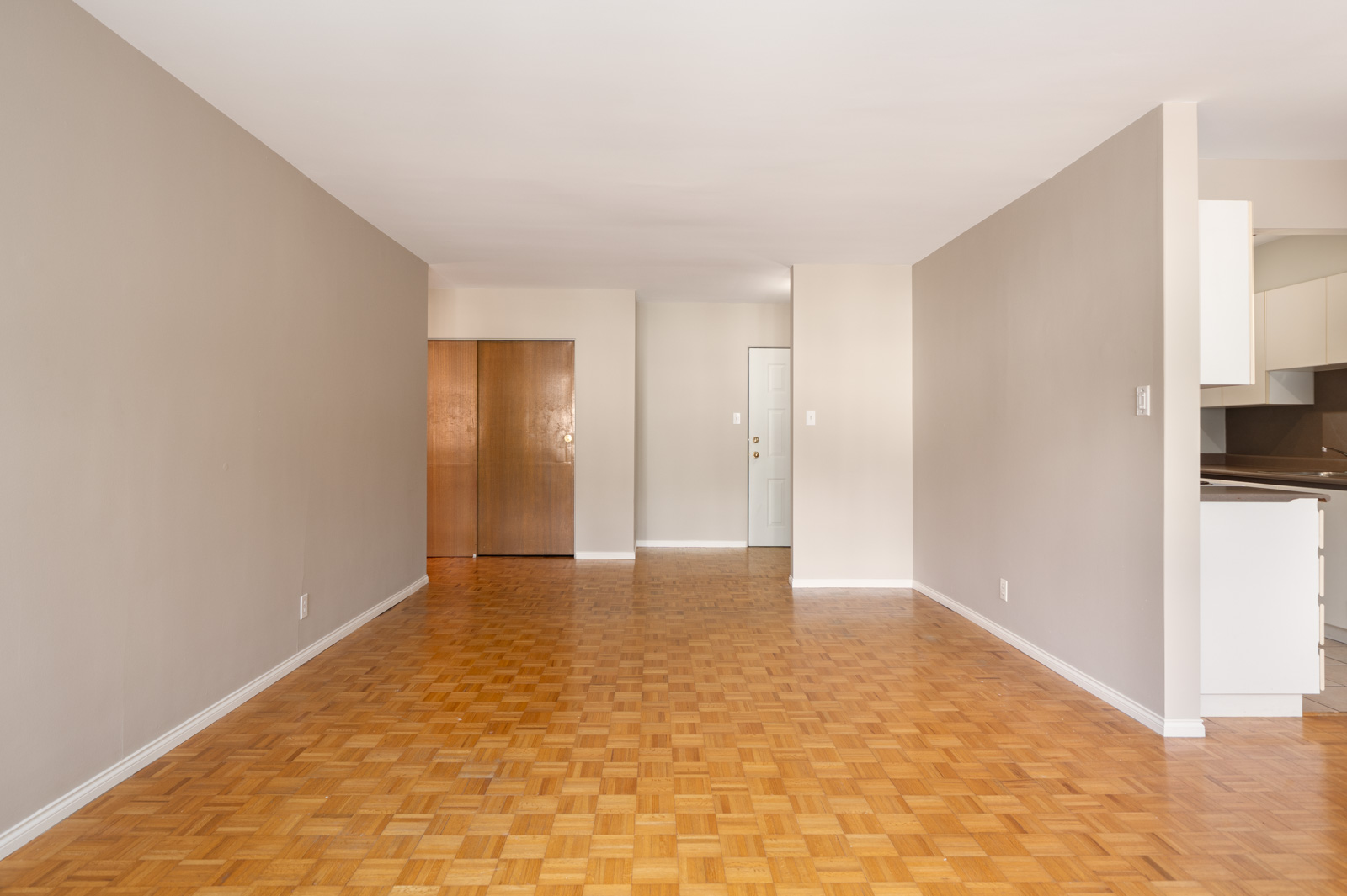Empty living room with light brown parquet flooring, beige walls, and a view of a closed wooden closet and a white entrance door in the background.