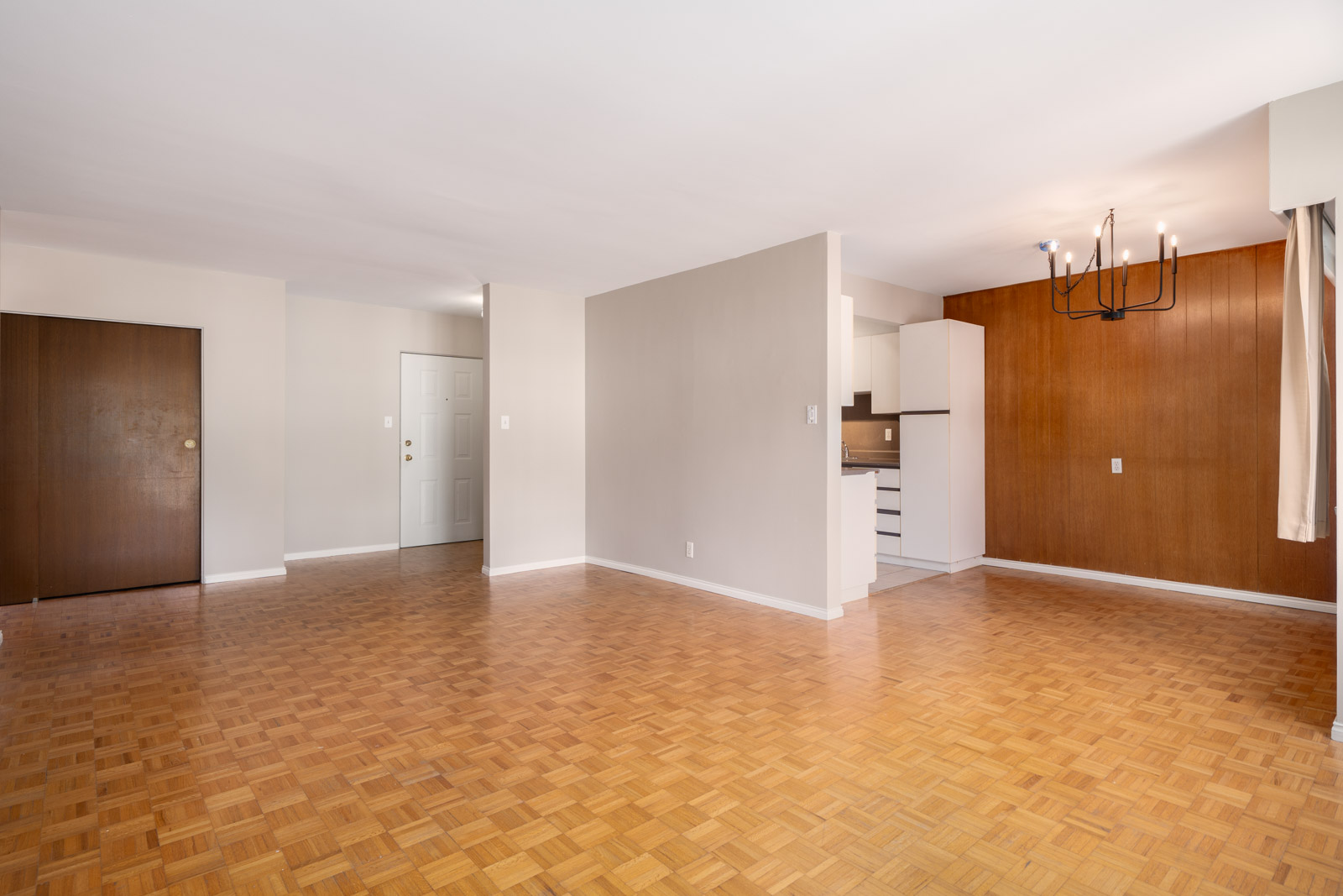 Unfurnished apartment interior with parquet flooring, white walls, wood-paneled accent wall, visible kitchen area, and closed doors.