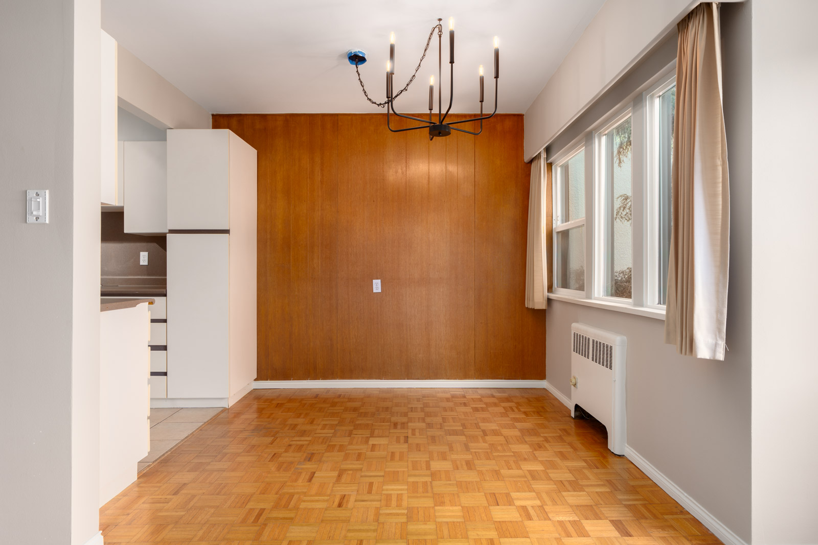 Empty dining area with parquet flooring, wood-paneled accent wall, modern chandelier, large windows with beige curtains, and adjacent kitchen and radiator.