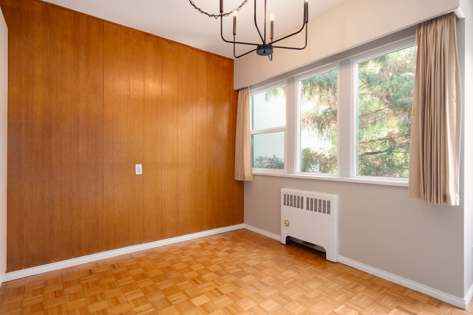 Empty room with wood-paneled wall, parquet floor, large window with beige curtains, radiator, and modern black chandelier.