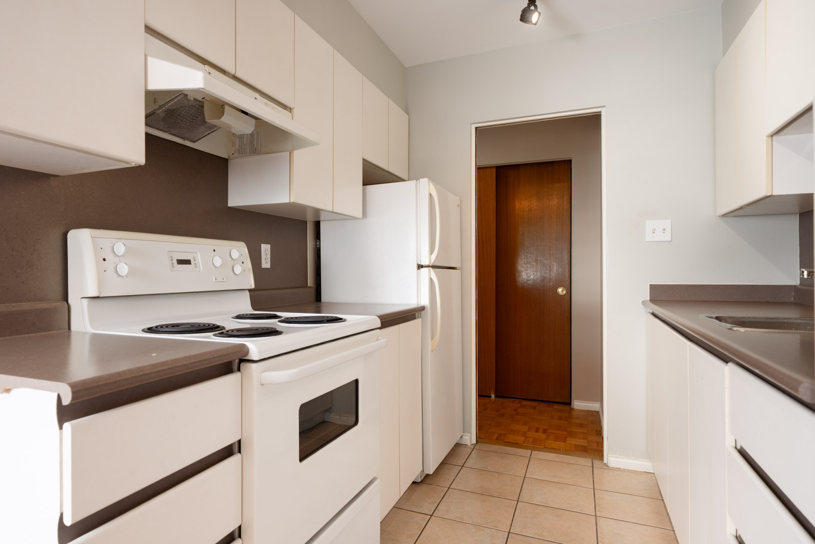 Narrow kitchen with white cabinets, white appliances including stove and refrigerator, brown countertops, and tile flooring.