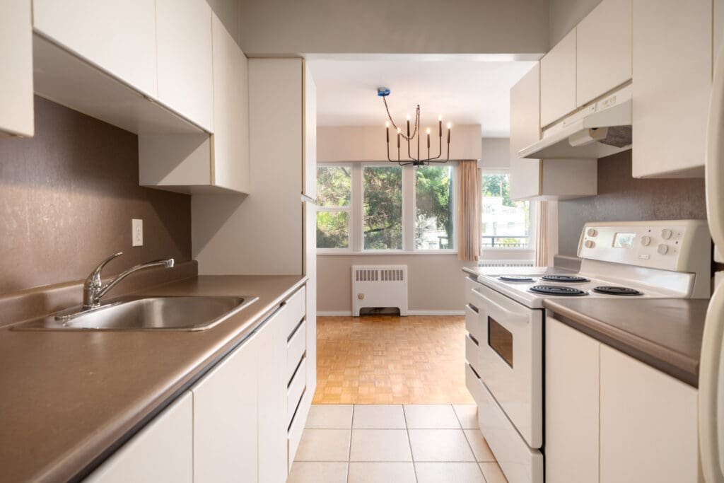 A narrow kitchen with white cabinets, brown countertops, electric stove, sink, and a view into a dining area with large windows and a modern chandelier.
