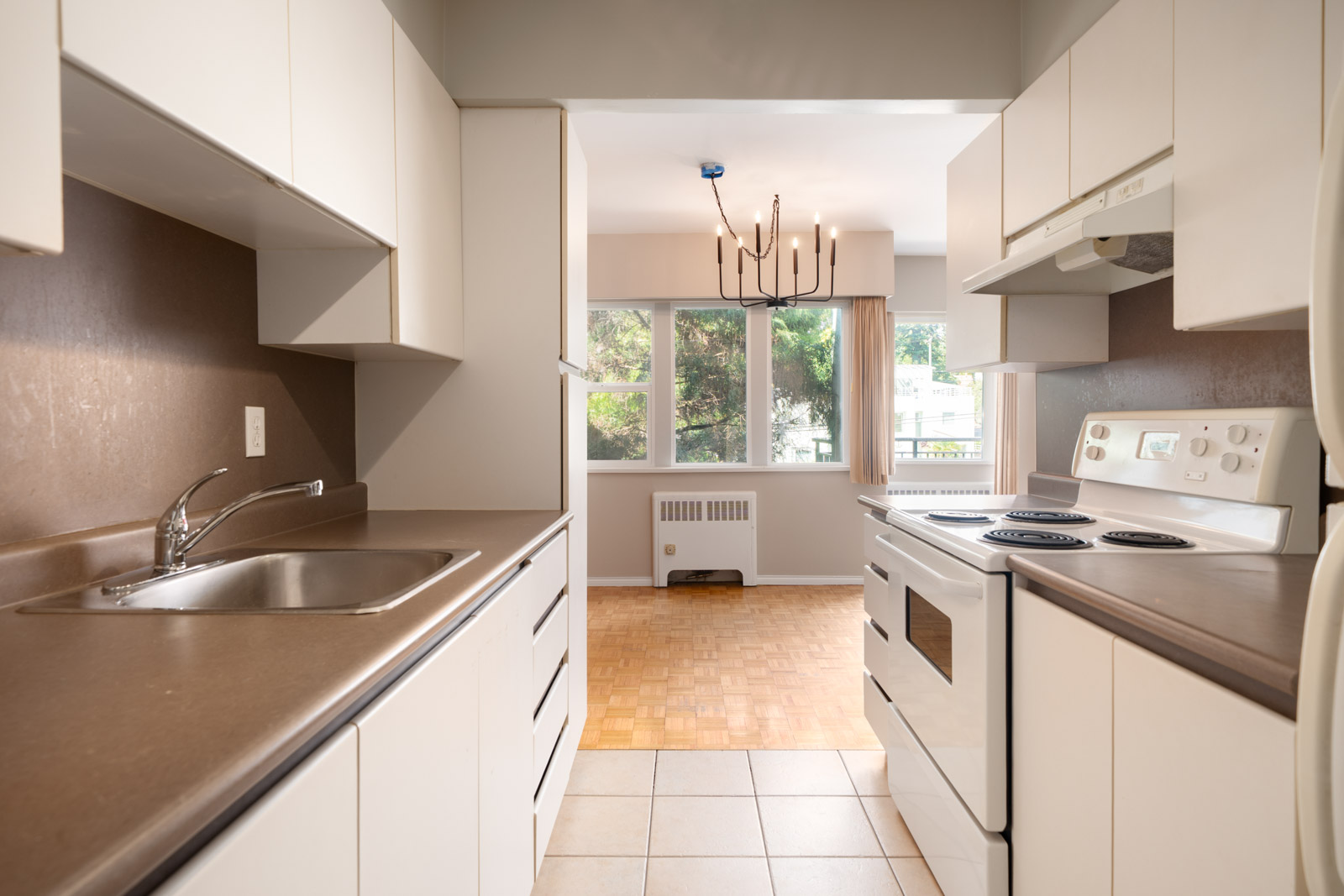 A narrow kitchen with white cabinets, brown countertops, electric stove, sink, and a view into a dining area with large windows and a modern chandelier.