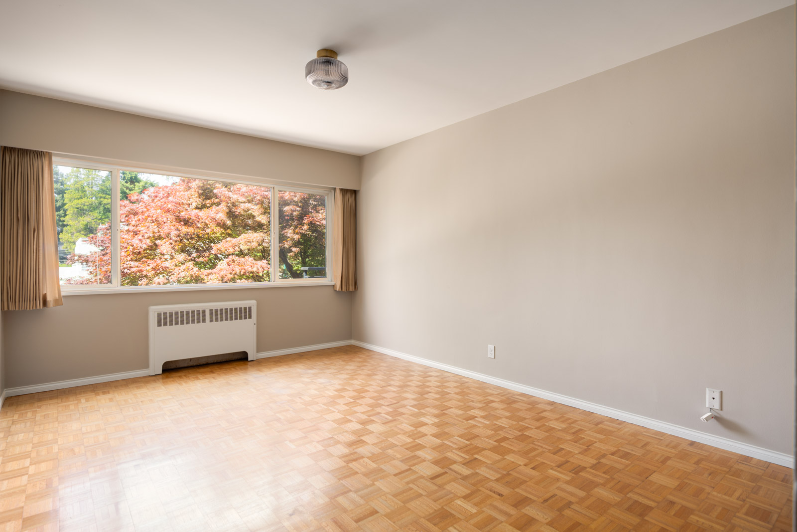 Empty room with light wood parquet flooring, beige walls, a large window with tan curtains, and a ceiling light fixture; trees visible outside.