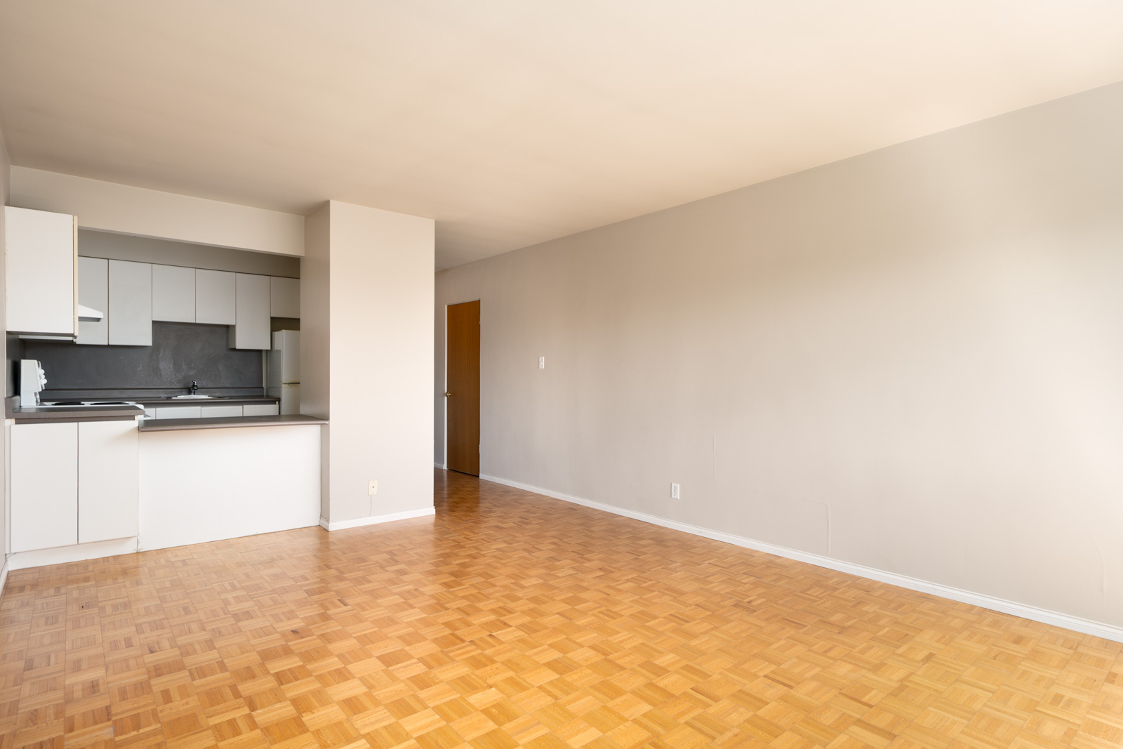 Unfurnished apartment with an open kitchen, white cabinets, parquet floors, and neutral-colored walls. A hallway and closed door are visible in the background.