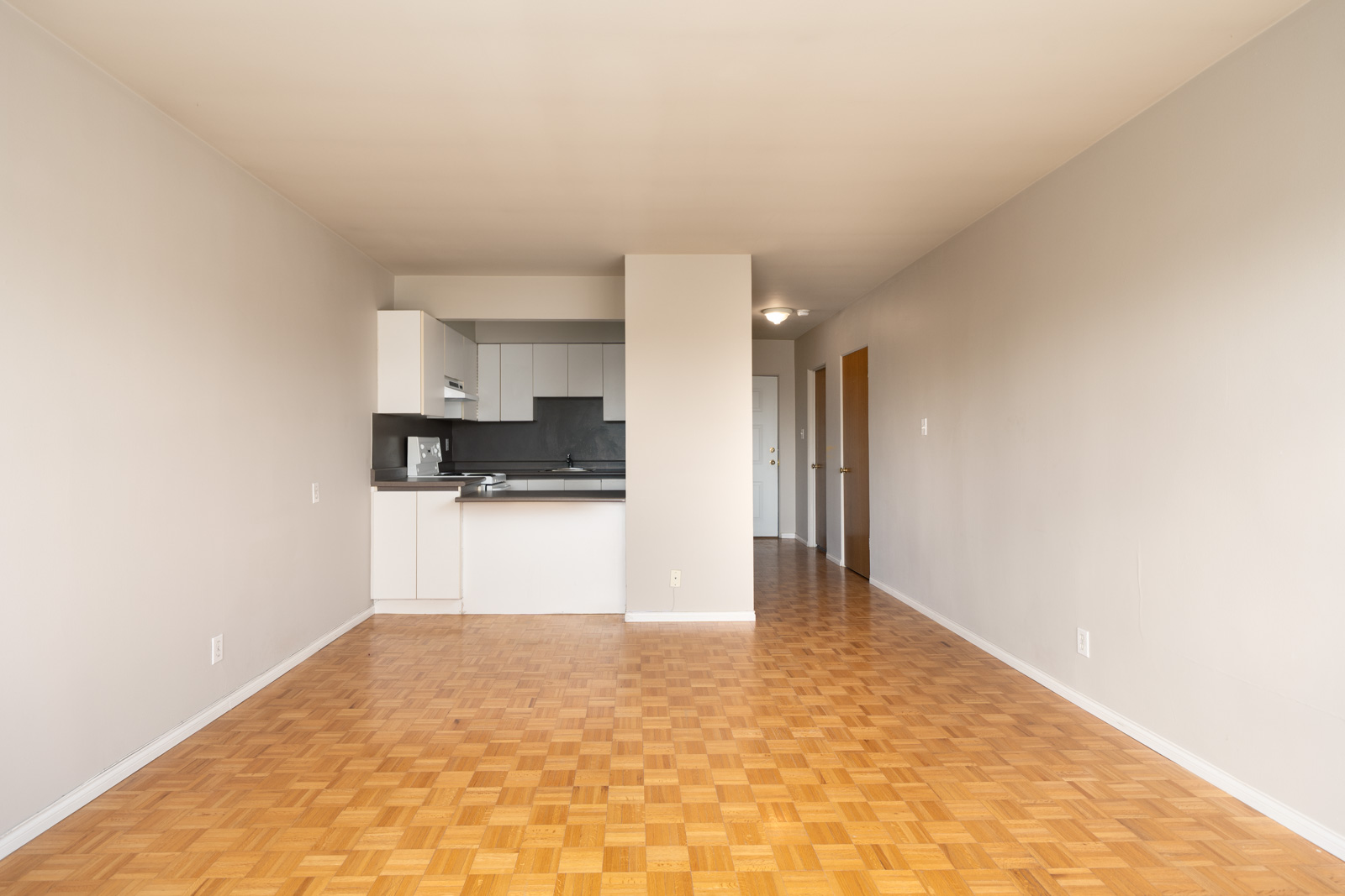 Empty apartment with parquet wood floors, white walls, a small open kitchen on the left, and a hallway leading to a door in the background.