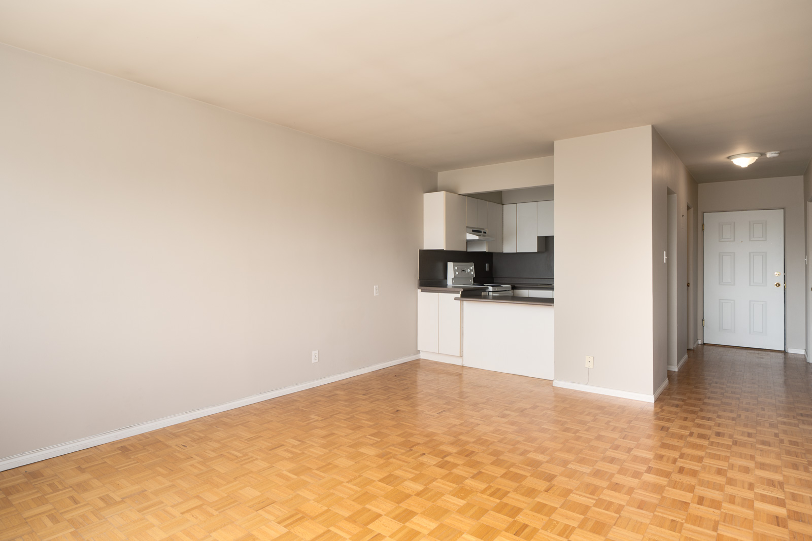 Empty apartment with light wood parquet floors, beige walls, a small white kitchen, and a hallway leading to a closed white entry door.