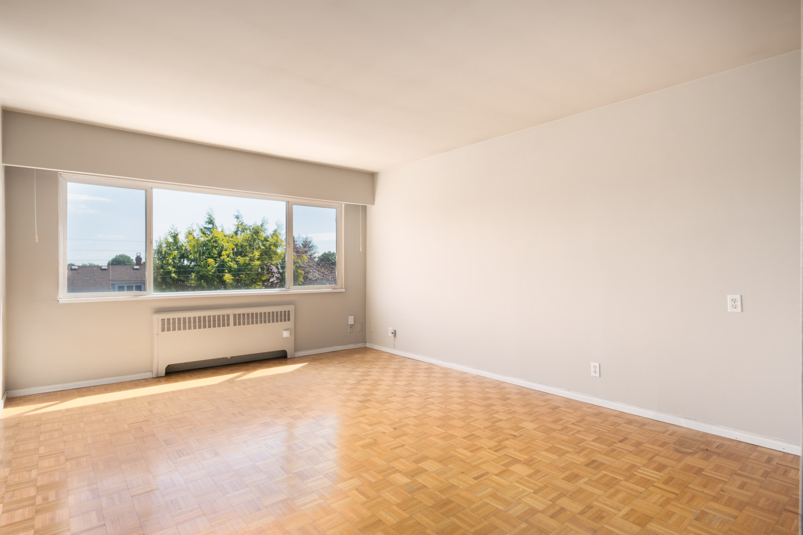 Empty room with parquet flooring, large window letting in natural light, white walls, and a baseboard heater under the window.