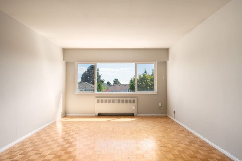 Empty room with light-colored walls, parquet flooring, a large window, radiator, and view of trees and sky outside.