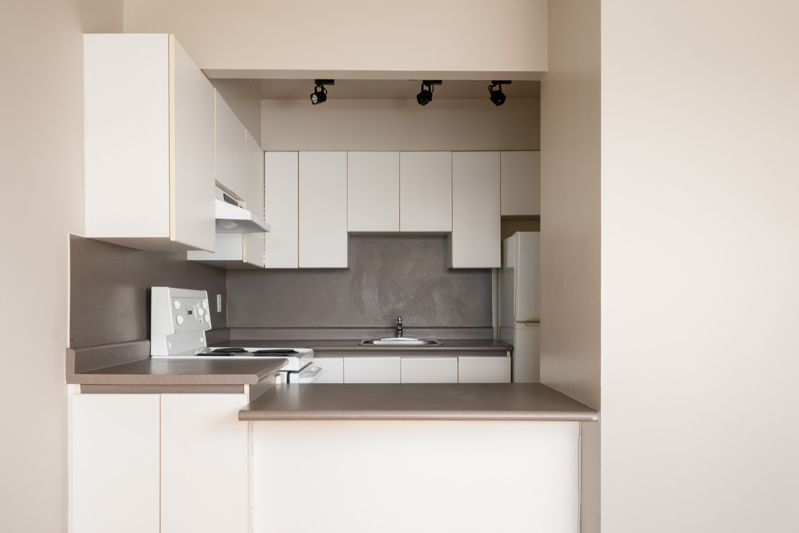 Minimalist kitchen with white cabinets, gray countertops, electric stove, sink, and overhead track lighting; neutral beige walls.