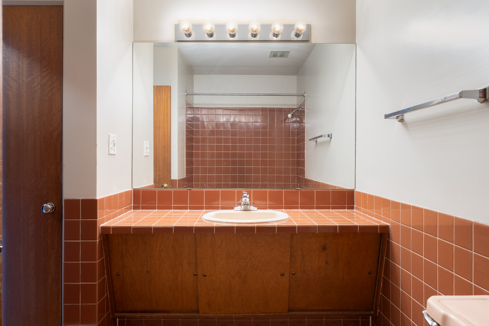 A bathroom with brown tiled countertop and backsplash, a white sink, large mirror, row of vanity lights, and a towel bar on the right wall.