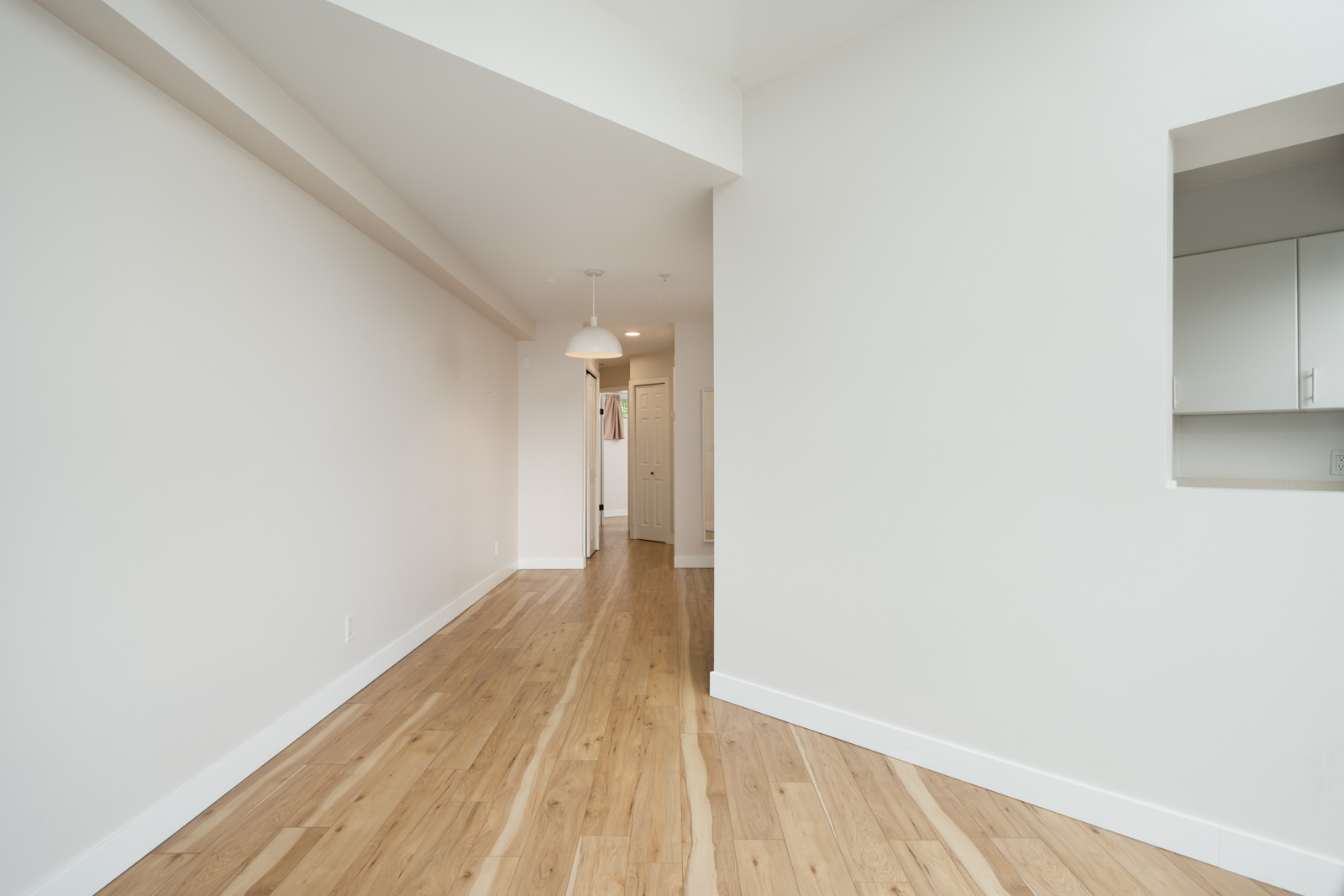 Empty room with light wood flooring, white walls, a ceiling light fixture, and a partial view of a kitchen through a wall opening.