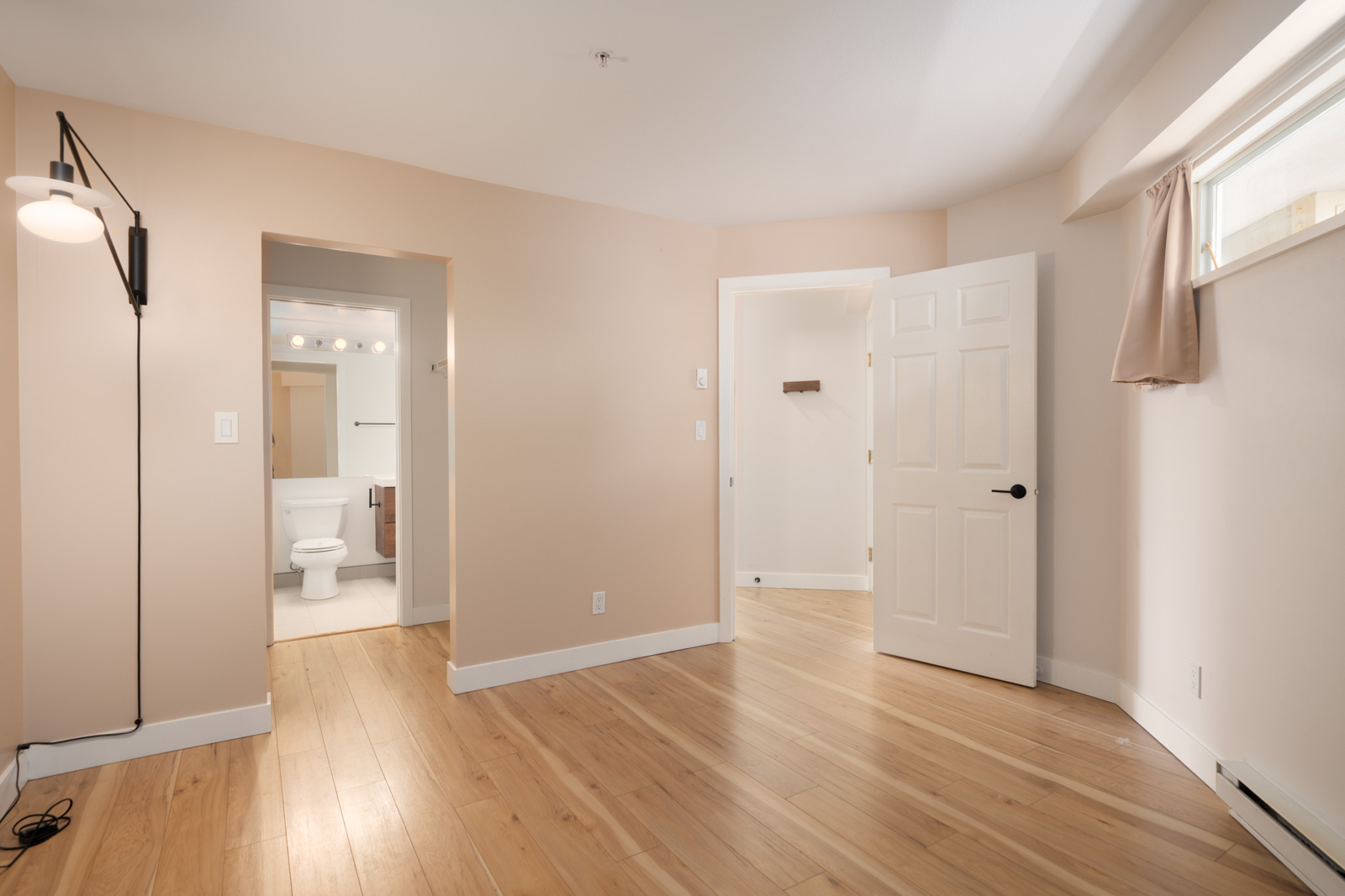 Empty beige bedroom with light wood flooring, open doorways to a bathroom and hallway, a wall-mounted lamp, and a small window with a curtain.
