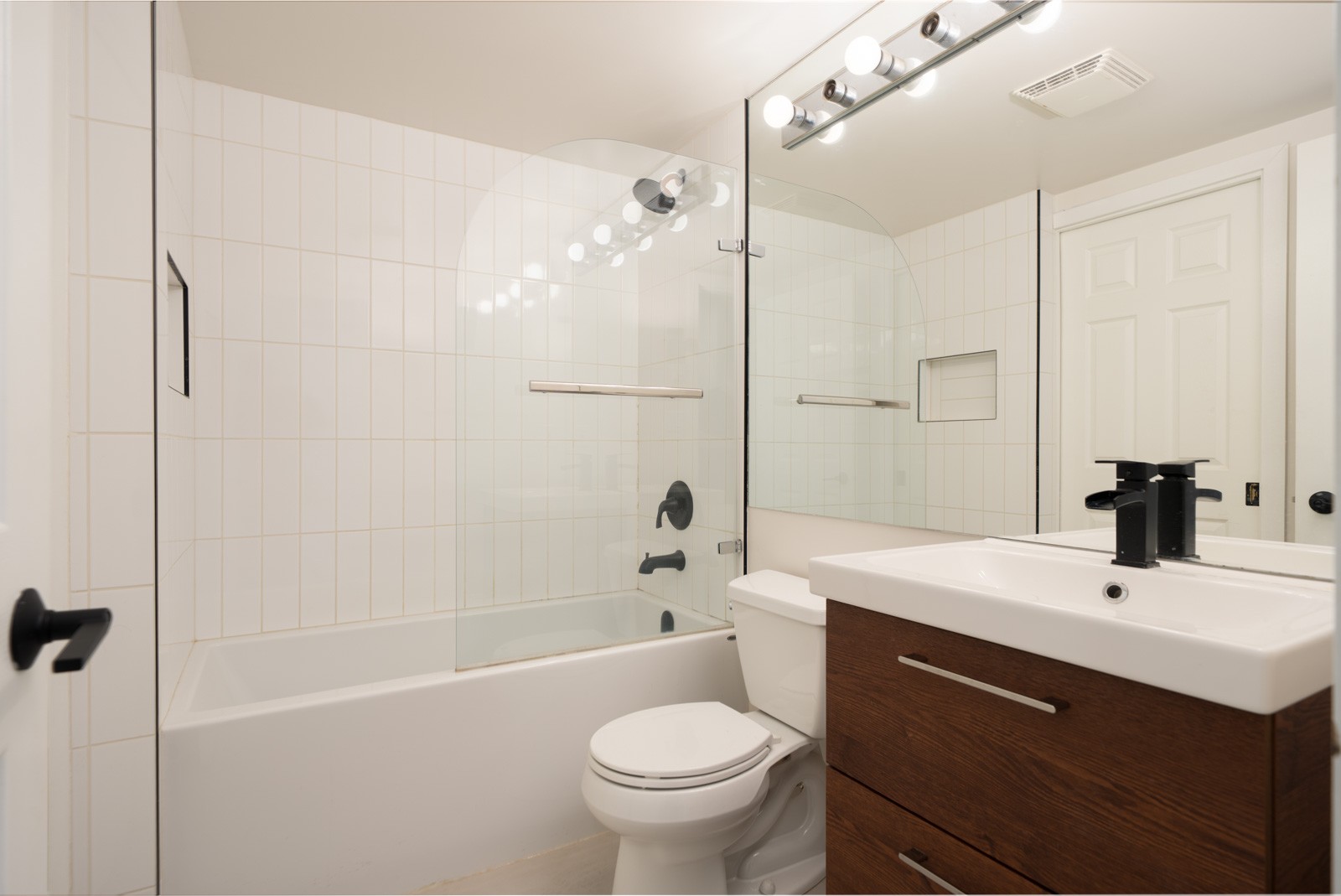 Modern bathroom with a bathtub and glass shower screen, white tile walls, a toilet, and a vanity with a rectangular sink and black fixtures.