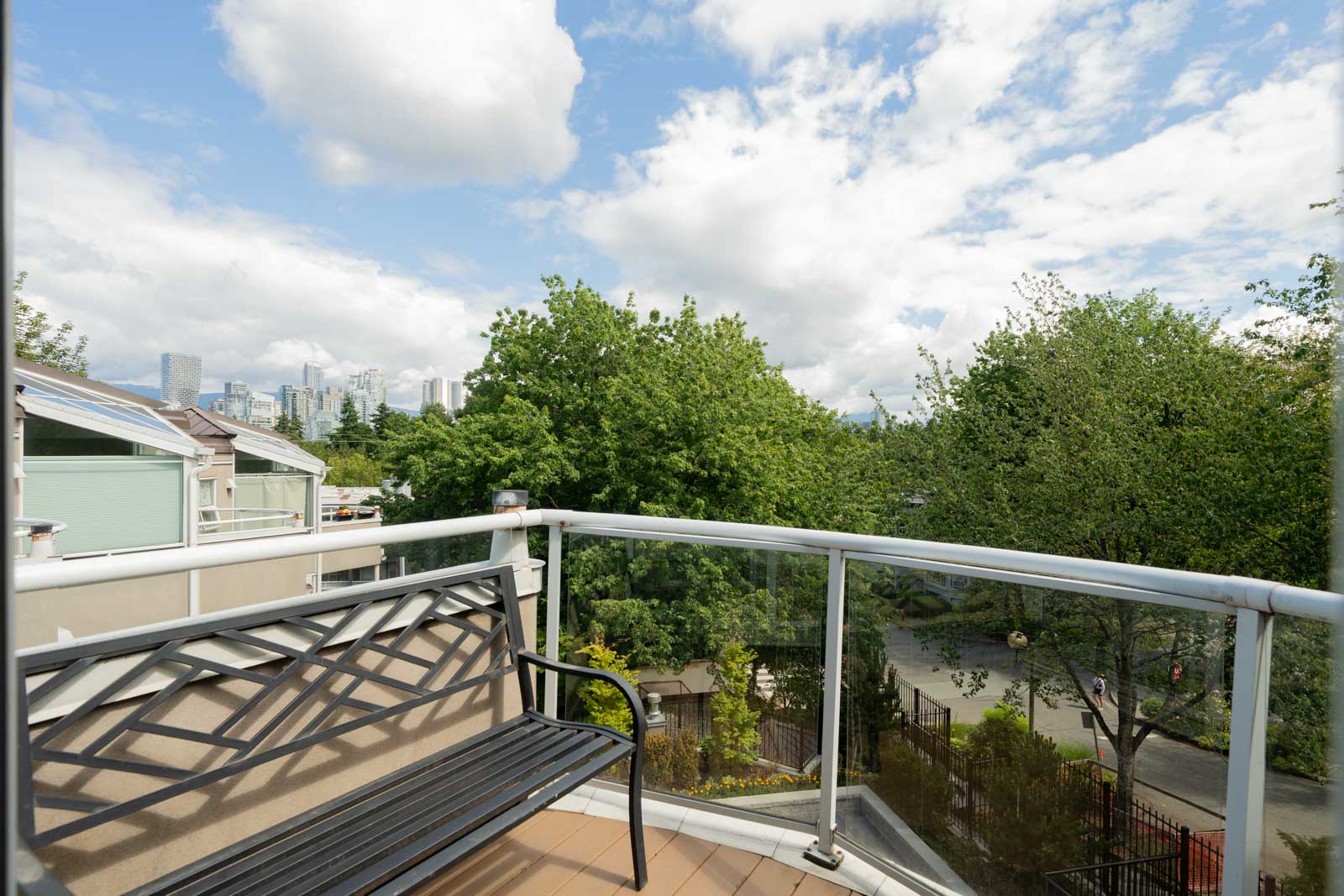 Balcony with a metal bench and glass railing overlooking trees, a park, and distant city buildings under a partly cloudy sky.