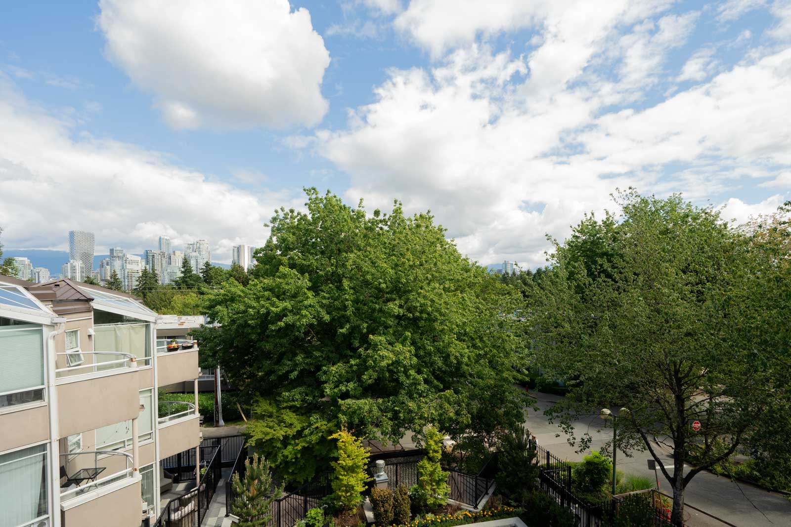 View of a residential area with apartment buildings, large green trees, and a distant city skyline under a partly cloudy sky.