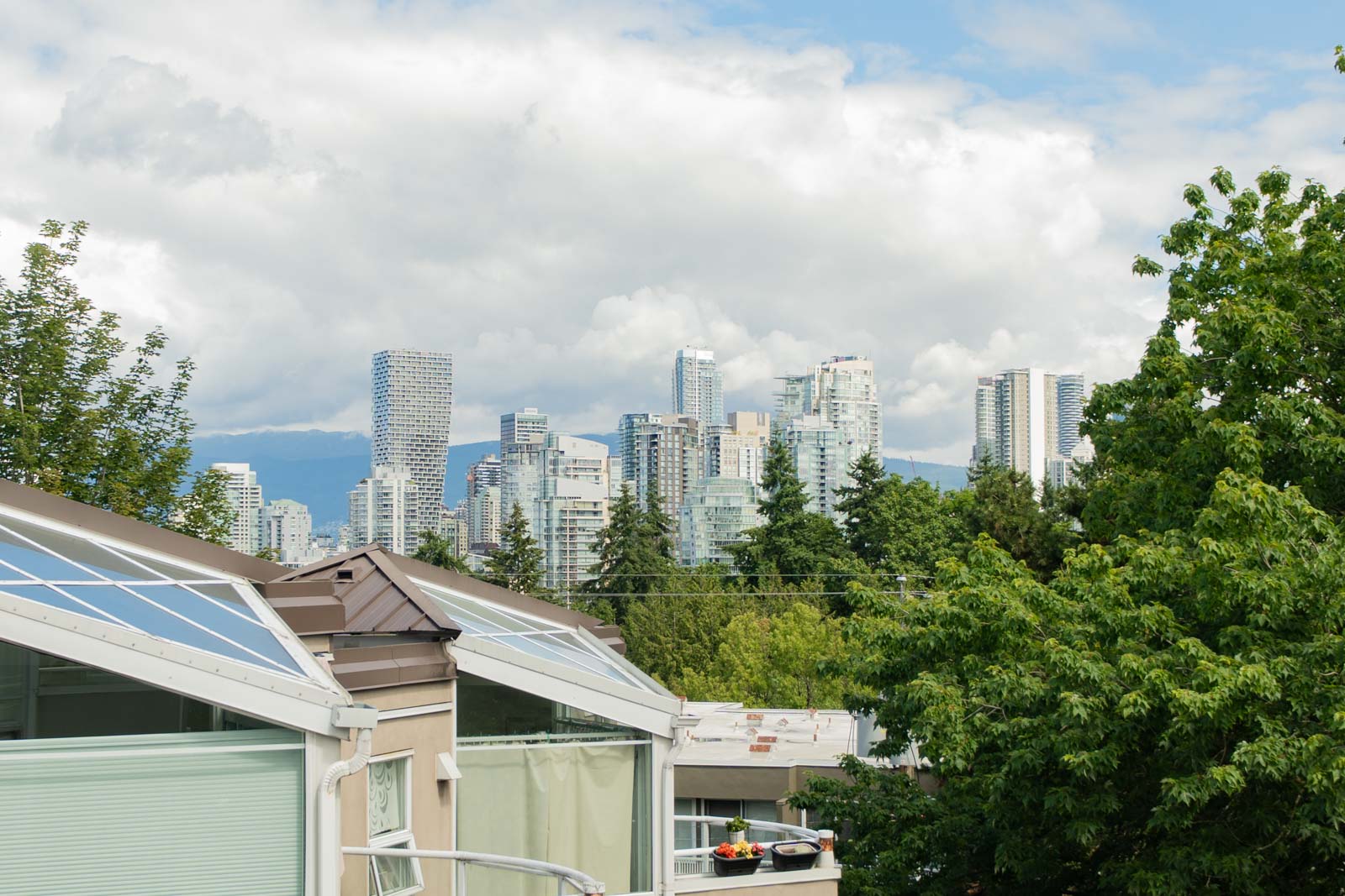 City skyline with tall modern buildings in the background, trees and residential rooftops with skylights in the foreground under a partly cloudy sky.