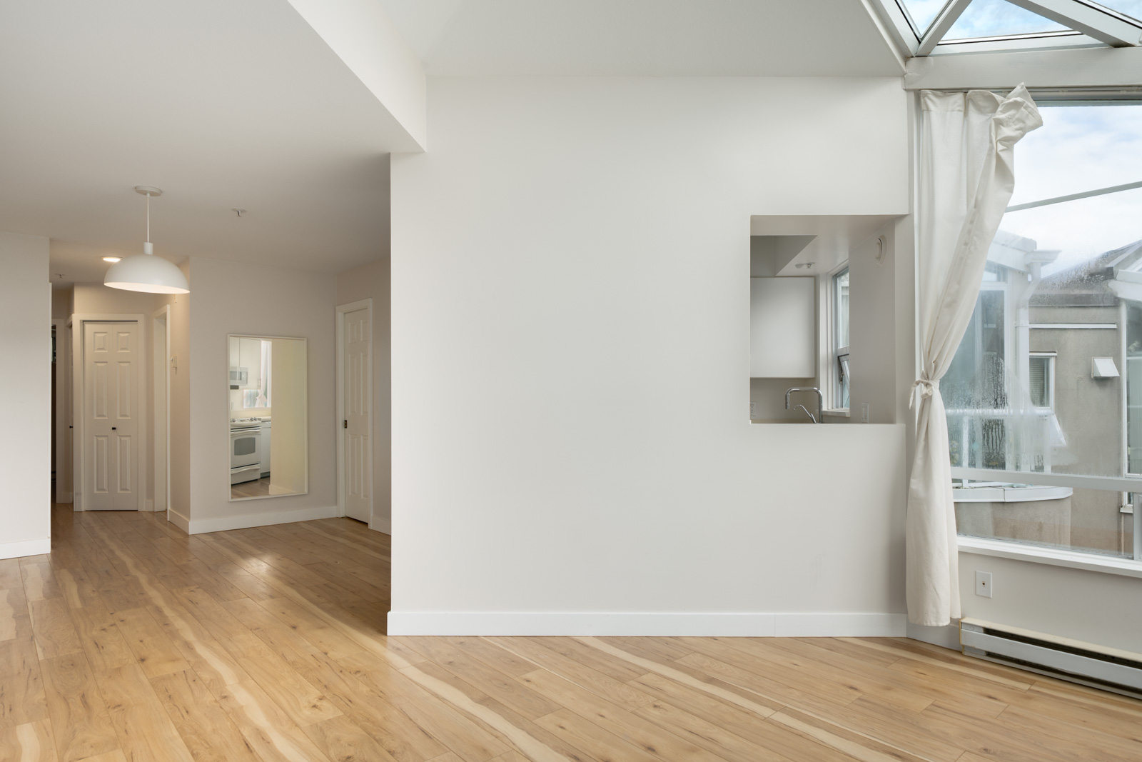 Bright, empty room with light wood floors, white walls, a small wall mirror, large window with curtain, and view into a kitchen through a doorway.