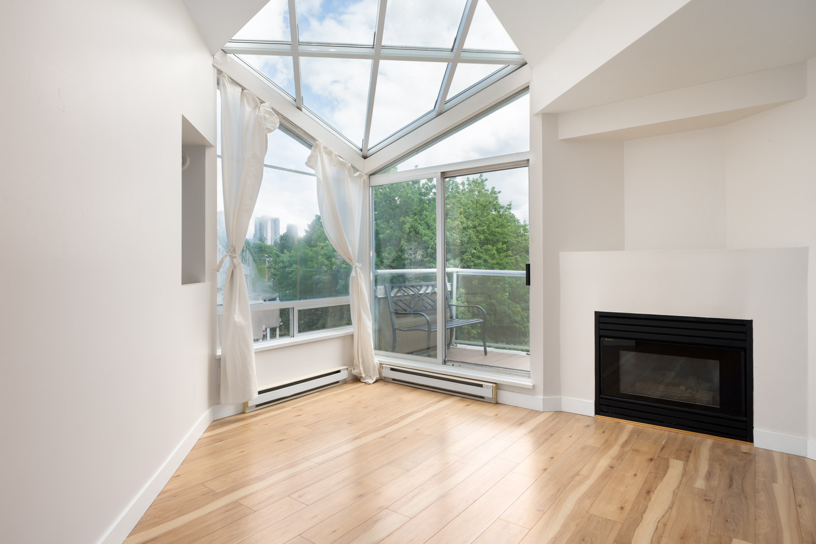 A bright, empty room with light wood flooring, a glass ceiling, large windows, white curtains, a fireplace, and a view of trees and buildings outside.