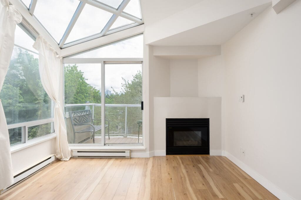Bright room with wood flooring, a modern fireplace, large windows, glass doors to a balcony, and a glass ceiling letting in natural light.