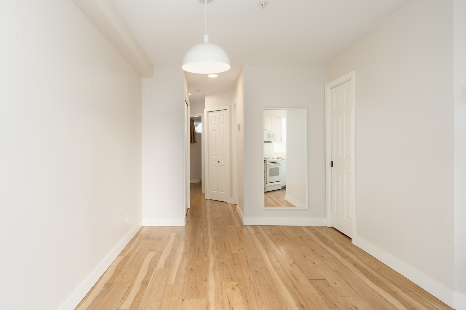 Empty room with light wood floors, white walls, a pendant light, a door, a large wall mirror, and a view into a kitchen with white appliances in the background.