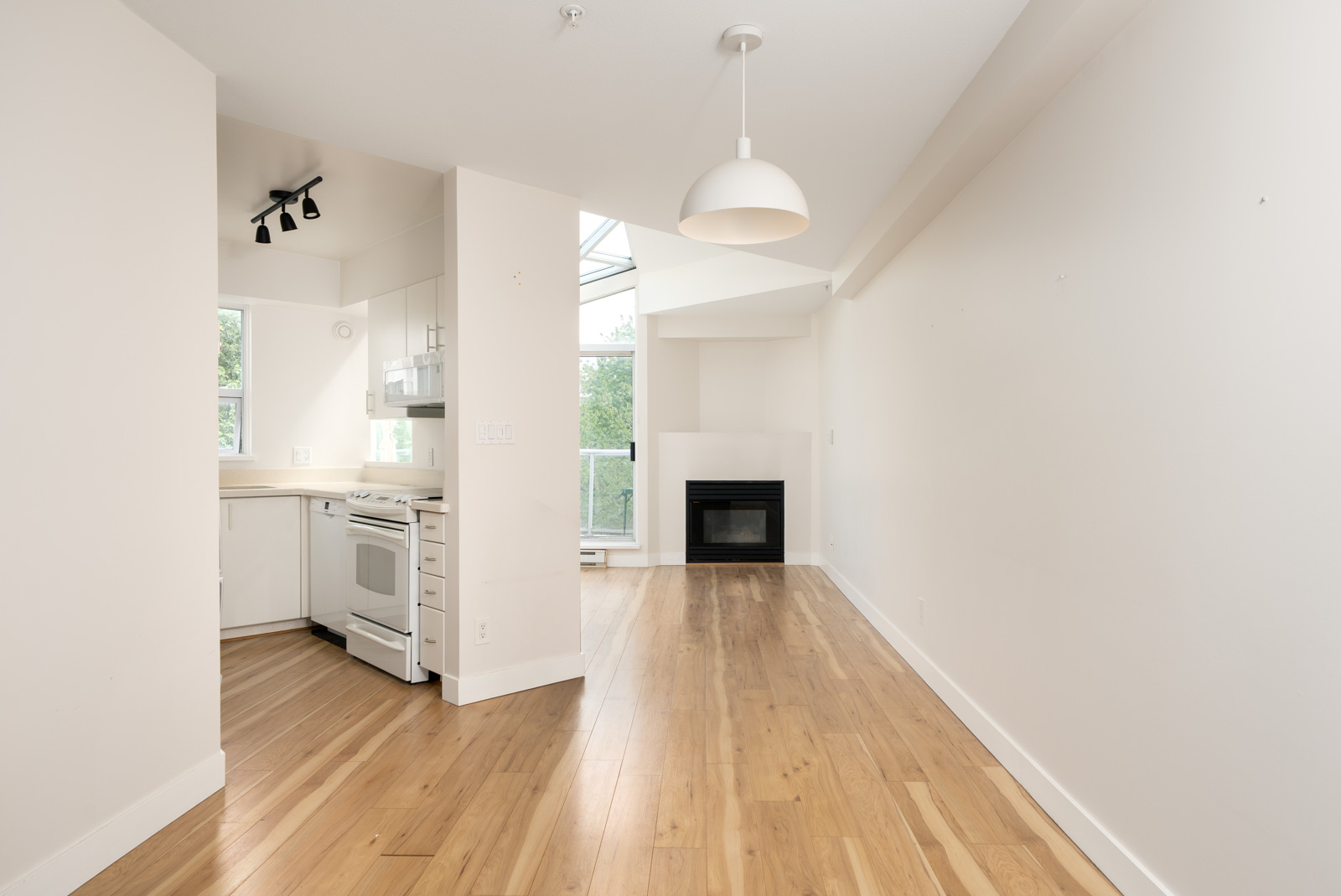 Bright, empty apartment with light wood floors, white walls, a small kitchen with white appliances, a fireplace, and large windows letting in natural light.