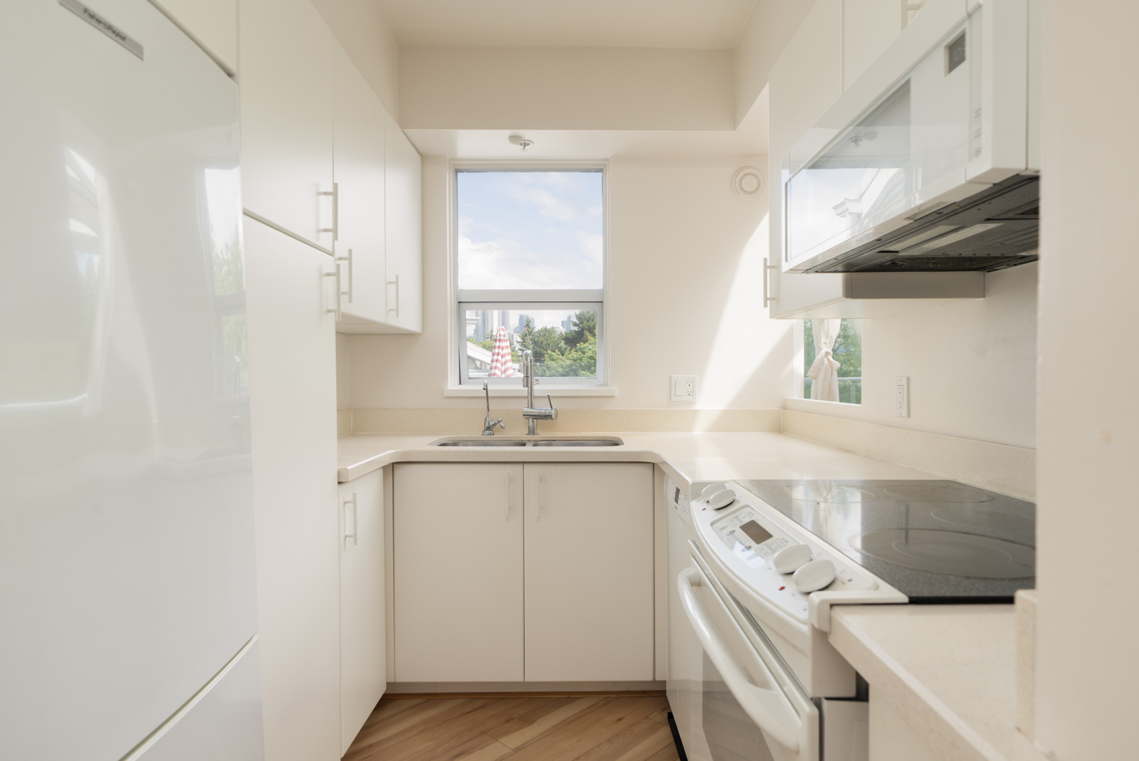 Small, modern kitchen with white cabinets, electric stove, microwave, refrigerator, and a window above the sink letting in natural light.
