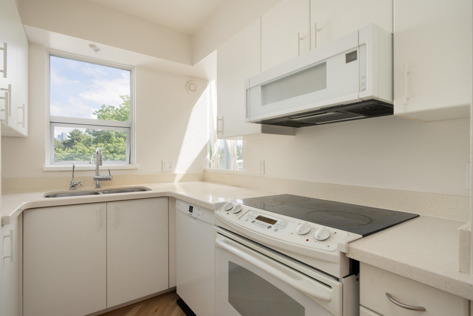 A modern white kitchen with an electric stove, built-in microwave, dishwasher, and a sink under a window with natural light.