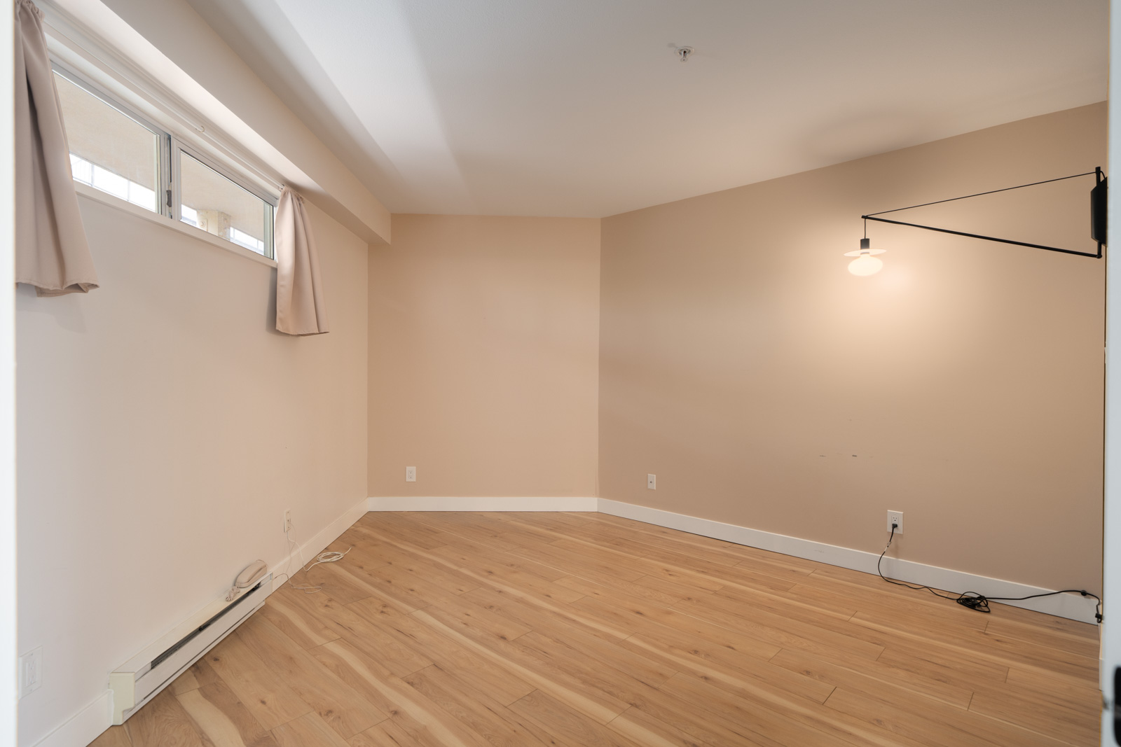 Empty room with light wood flooring, beige walls, a high window with short curtains, a wall-mounted lamp, and exposed electrical cords along the baseboard.