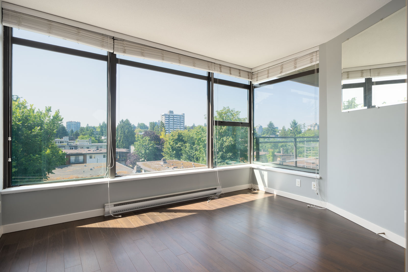 Bright corner room at 1068 W Broadway, Vancouver, featuring wood floors, tall windows, and views of trees and nearby buildings.