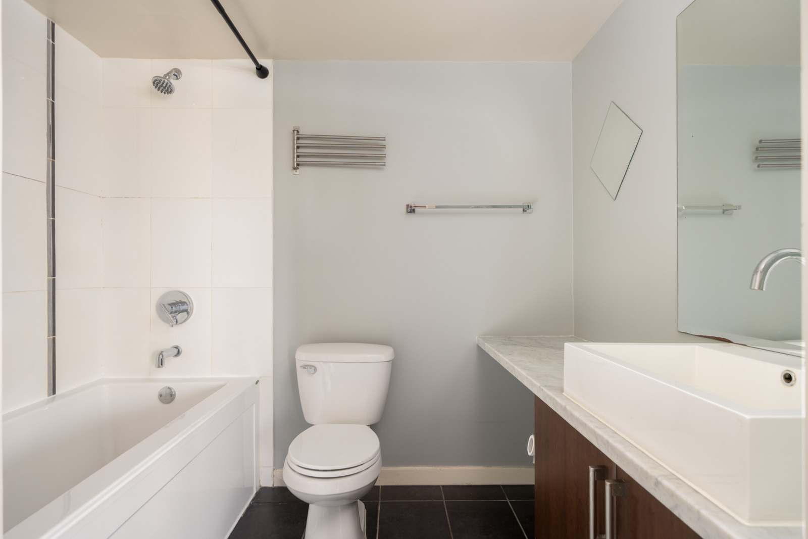 Contemporary bathroom at 1068 W Broadway, Vancouver with tub, toilet, wall sink, large mirror, and towel racks on gray walls.