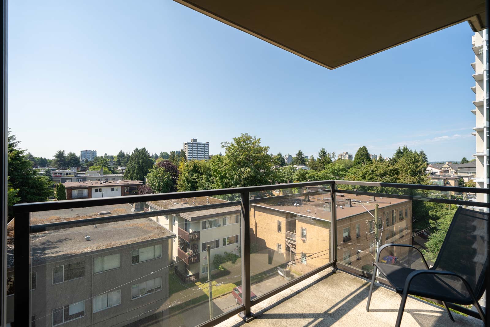 Balcony with glass railing overlooks low-rise buildings, trees, and Vancouver skyline with 1068 W Broadway under blue sky.