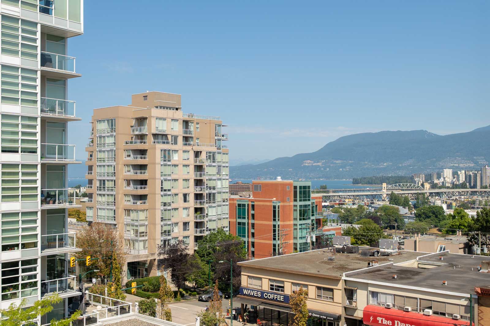 Modern apartment buildings, a coffee shop, and distant mountains under a blue sky in Vancouver near 1068 W Broadway.