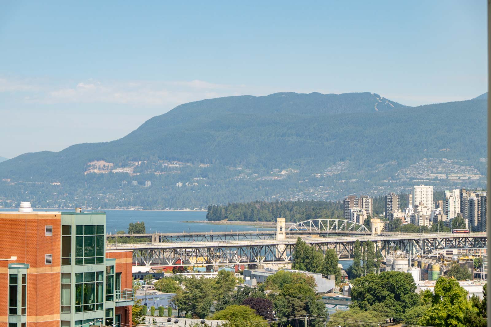 Modern Vancouver cityscape with unique buildings by 1068 W Broadway, bridge and water in front, forested mountains behind.