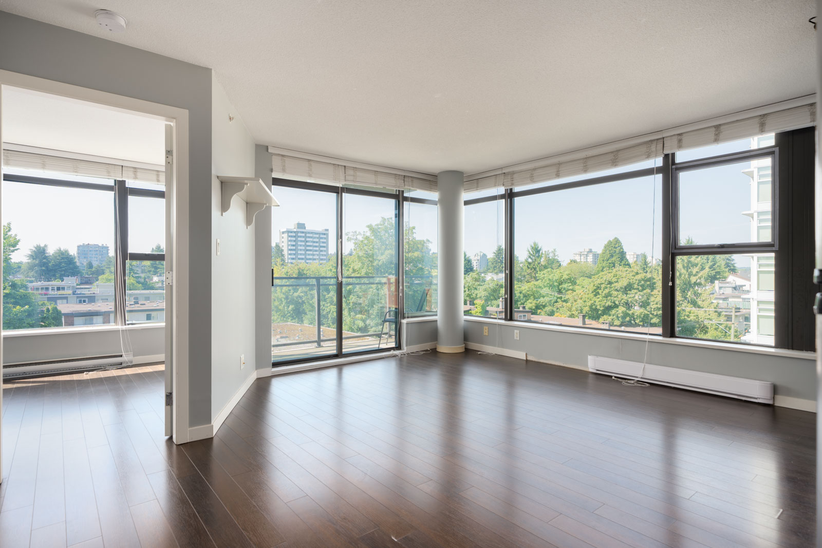 Sunny, unfurnished living room with large windows, dark hardwood floors, and views of trees and city buildings in Vancouver.
