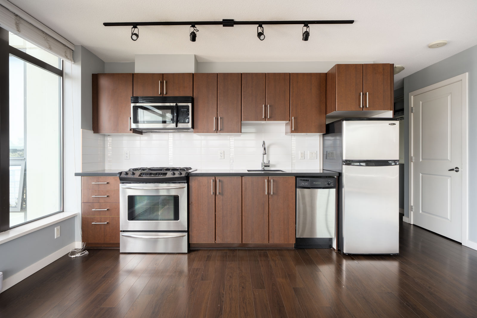 Modern kitchen at 1068 W Broadway, Vancouver with stainless appliances, dark wood cabinetry, white tile backsplash, and large window.