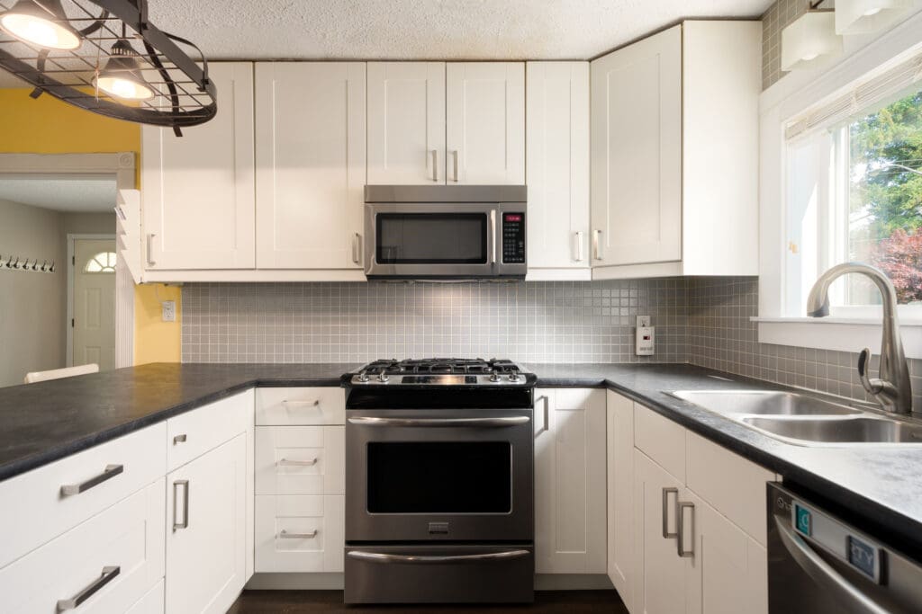 Modern kitchen with white cabinets, gray tile backsplash, stainless steel stove and microwave, double sink, and a window letting in natural light.