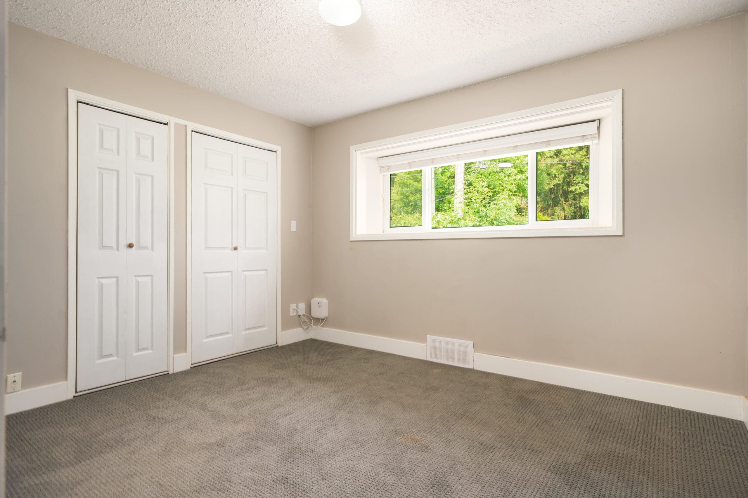 Empty room with beige walls, a grey carpet, two white double-door closets, and a large window showing greenery outside.