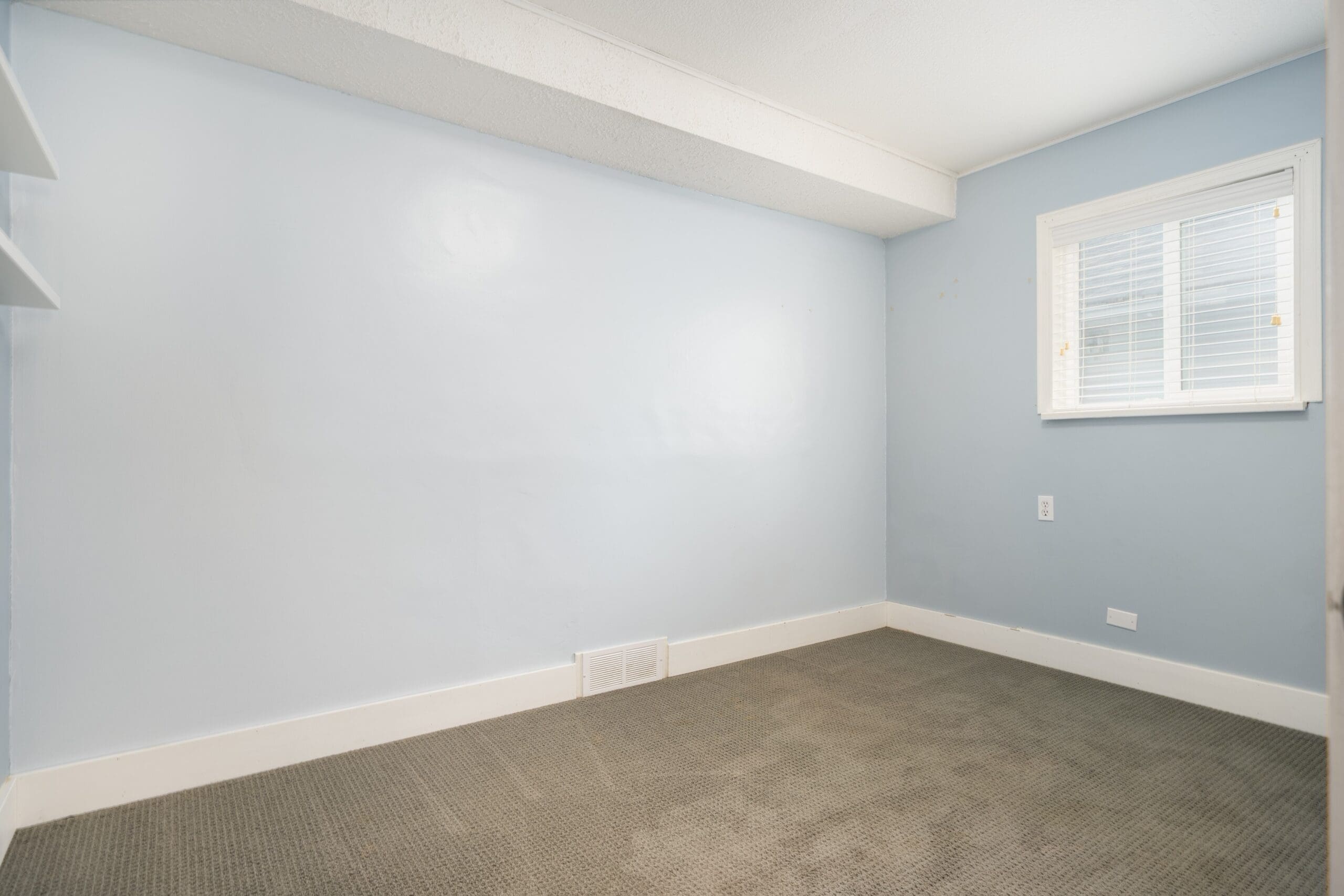 Empty room with light blue walls, a small window with blinds, gray carpet, white trim, and a wall vent near the floor.