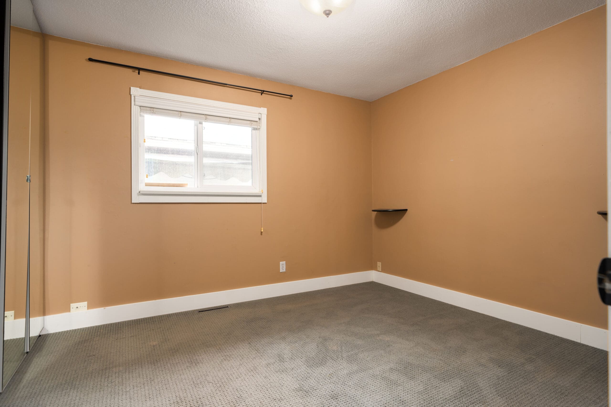 Empty room with beige walls, grey carpet, a window with white blinds, a ceiling light, a wall shelf, and a mirrored closet on the left.